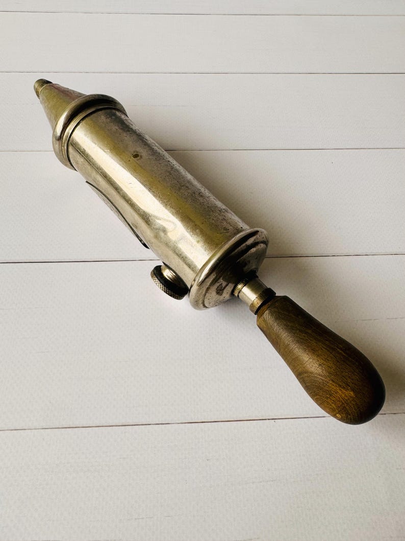 Vintage Perolin grease gun shown at an angle, antique tool with worn metal surface and wooden handle, ideal for industrial decor or man cave decor