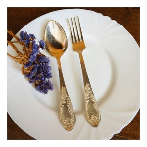 Pode incluir: Uma colher e um garfo de prata com desenhos ornamentados em um prato branco com um ramo de lavanda seca.