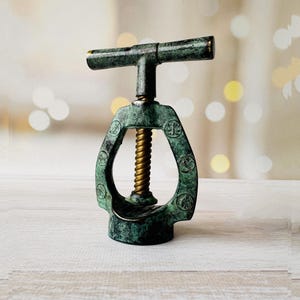 May include: Antique bronze-colored corkscrew with a T-shaped handle and a spiral screw. The corkscrew has a weathered, aged appearance with a green patina and gold accents. The design includes decorative circular cutouts.