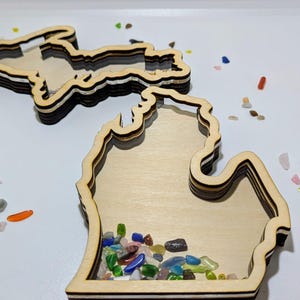 May include: Two wooden trays in the shape of the state of Michigan. One tray is filled with colorful glass stones. The trays are made of layered wood with a natural finish. The trays are on a white surface.