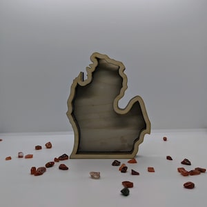 May include: A wooden cutout of the state of Michigan, standing on a white surface with small red and brown stones scattered around it.