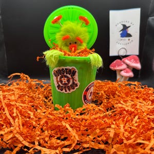May include: A whimsical green trash can with a fluffy green creature inside, surrounded by orange paper shreds. The trash can has a lid and stickers with the text "Houston Astros". A small card and mushroom figurines are in the background.