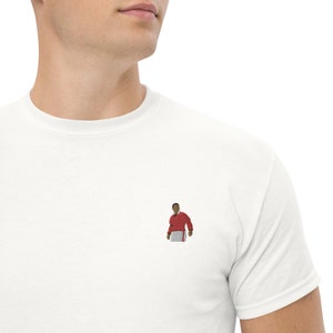 Puede incluir: Camiseta blanca con una pequeña imagen bordada de una persona que lleva una camisa roja y un pantalón blanco.