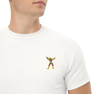 Puede incluir: Camiseta blanca con un pequeño diseño bordado de un luchador en amarillo, marrón y rojo. El luchador está representado en una pose dinámica con los brazos levantados. La camiseta está hecha de un material suave y cómodo.
