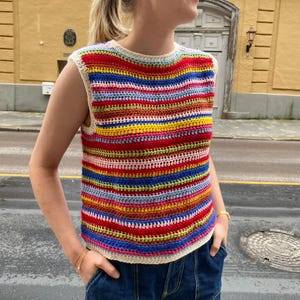 Op de afbeelding: Een mouwloos gebreid topje met een regenboogkleurig gestreept patroon. Het topje is gemaakt van wit garen en heeft een ronde hals.