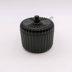 May include: A black, ribbed glass sugar bowl with a matching lid and a small silver spoon. The bowl has a round shape with vertical grooves and a small knob on the lid. The spoon rests on the edge of the bowl.