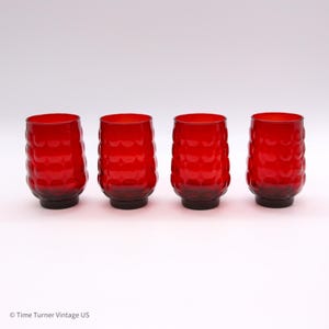 May include: A set of four cylindrical red glass tumblers, each featuring a repeating embossed circular pattern across the exterior surface. The glasses have a smooth rim and a slightly tapered base, standing upright in a row against a plain background.