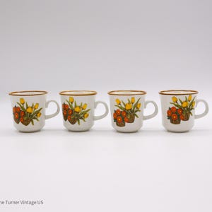 May include: Four white ceramic mugs with a floral design. Each mug features a basket of orange and yellow flowers with a brown rim and handle. The mugs are arranged in a row against a white background.