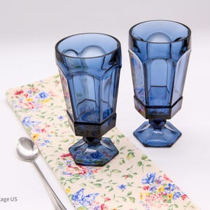 May include: Two cobalt blue glass goblets with a faceted, paneled design and a hexagonal base. The glasses are positioned on a cream-colored fabric featuring a colorful floral and butterfly pattern, accompanied by a single silver-toned metal spoon.