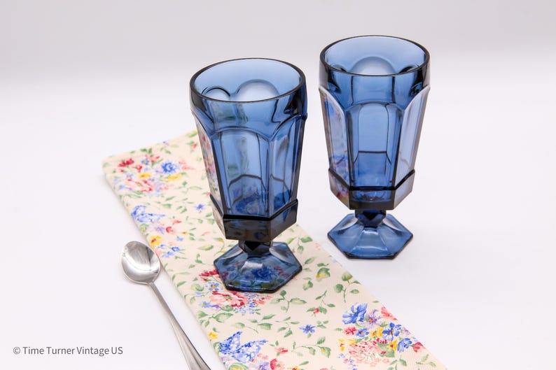 May include: Two cobalt blue glass goblets with a faceted, paneled design and a hexagonal base. The glasses are positioned on a cream-colored fabric featuring a colorful floral and butterfly pattern, accompanied by a single silver-toned metal spoon.