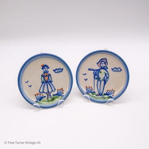 May include: Two small, round ceramic plates with a blue rim and hand-painted figures. One plate depicts a person in a dress, the other a person with a flute. Both plates have blue accents and are on clear stands.