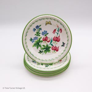 May include: A stack of white ceramic bowls with a green rim and floral design. The bowls feature red and blue flowers, green leaves, and butterflies. The bowls are stacked on top of each other.