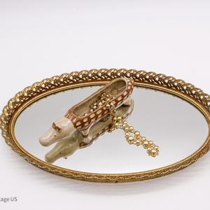 May include: A gold oval tray with a mirrored surface and a decorative, brown and white ceramic animal figurine inside. The figurine is shaped like a crocodile or alligator and has a small hole in its back, suggesting it may be a trinket dish.