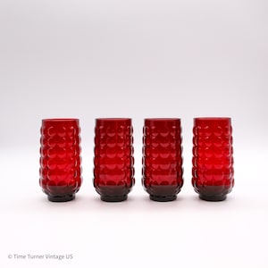 May include: A set of four cylindrical drinking glasses in a deep red color. Each glass features a repeating pattern of raised circular bumps covering the exterior surface, creating a textured finish. The glasses have a flat base and open top.