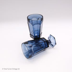 May include: Two translucent blue glass goblets with a faceted, hexagonal design. One goblet stands upright while the other rests horizontally on its side, showcasing the clear, deep blue color and the geometric structure of the glass stems.
