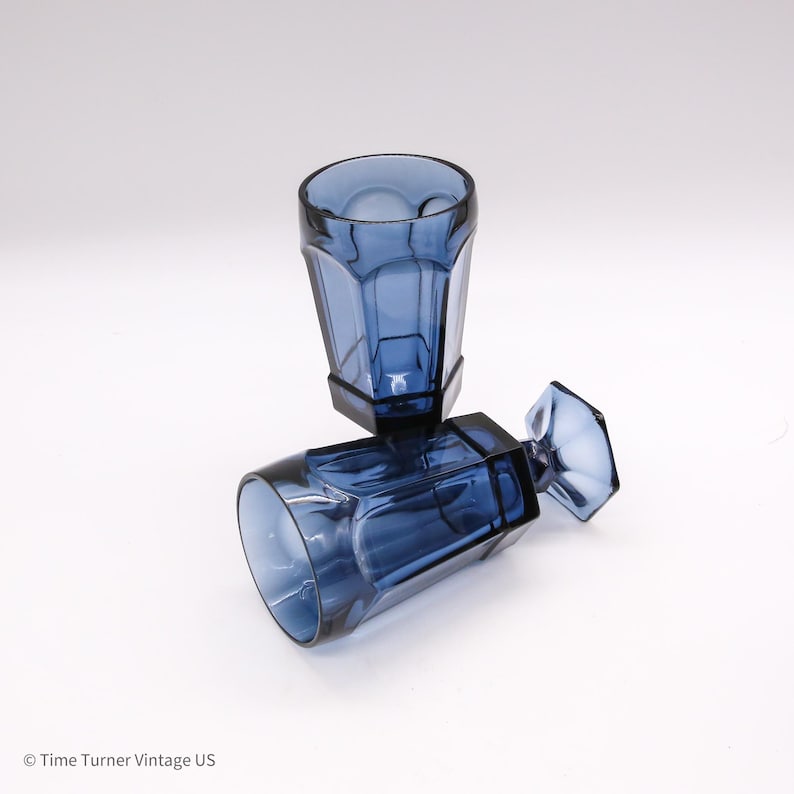 May include: Two translucent blue glass goblets with a faceted, hexagonal design. One goblet stands upright while the other rests horizontally on its side, showcasing the clear, deep blue color and the geometric structure of the glass stems.