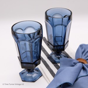 May include: Two blue glass goblets with a faceted, hexagonal design on the bowl and base. The glasses feature a smooth, rounded rim and a sturdy stem. A blue cloth napkin with a wooden ring is positioned next to the glassware.