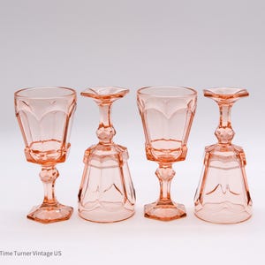 May include: Four vintage pink glass goblets with a decorative design. The goblets have a faceted bowl, a detailed stem, and a square base. The glasses are arranged in a row against a white background. The goblets are approximately 6 inches tall.