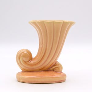 May include: A peach-colored ceramic cornucopia vase featuring a glossy finish and vertical fluted ridges. The vessel has a curved, horn-like shape that tapers toward a wide, flared opening at the top and rests on a solid, flat oval base.