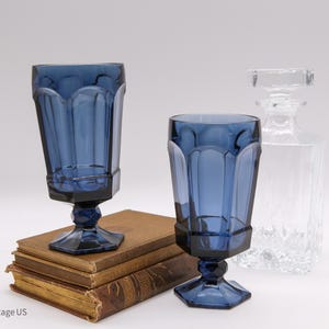 May include: Two cobalt blue glass goblets with a faceted, paneled design and hexagonal bases, accompanied by a clear glass decanter with a square stopper, all positioned near a stack of three vintage books with worn, tan-colored covers.