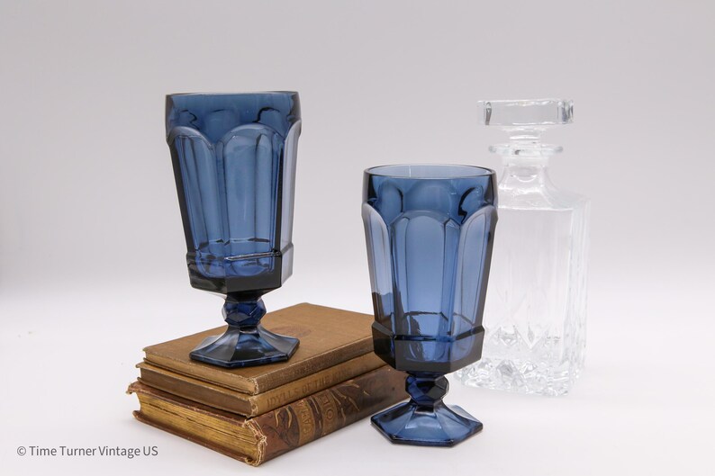 May include: Two cobalt blue glass goblets with a faceted, paneled design and hexagonal bases, accompanied by a clear glass decanter with a square stopper, all positioned near a stack of three vintage books with worn, tan-colored covers.