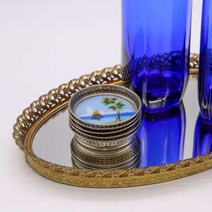 May include: A set of silver coasters with a blue and white painted design of a sailboat and palm trees on a mirrored oval tray with a gold metal frame.