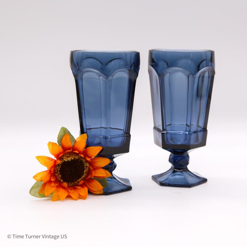 May include: Two tall, dark blue glass goblets with a faceted, paneled design and a hexagonal base. A single artificial sunflower with orange and yellow petals and a dark brown center is positioned in front of the left glass goblet.