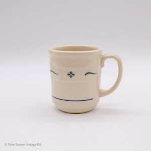 May include: Cream-colored ceramic mug with a curved handle. The mug features a blue floral design and horizontal blue lines. The mug is a classic design, suitable for coffee or tea.