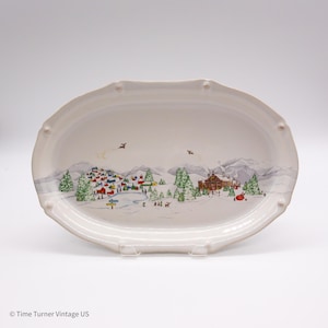 May include: An oval white ceramic serving platter featuring a detailed winter village scene with snow-covered mountains, evergreen trees, small houses, and figures. The rim has a scalloped edge with small raised decorative dots.