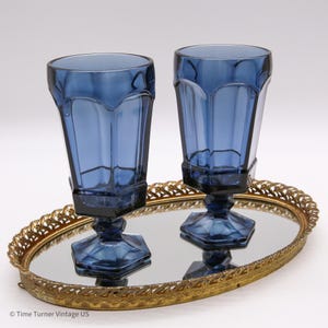 May include: Two tall, dark blue glass goblets with a faceted, paneled design and hexagonal bases. The glasses are positioned on an oval, gold-toned metal vanity tray with a mirrored surface and an intricate, openwork filigree border edge.