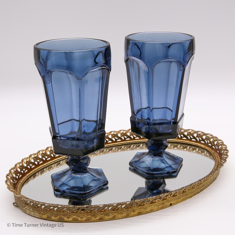 May include: Two tall, dark blue glass goblets with a faceted, paneled design and hexagonal bases. The glasses are positioned on an oval, gold-toned metal vanity tray with a mirrored surface and an intricate, openwork filigree border edge.
