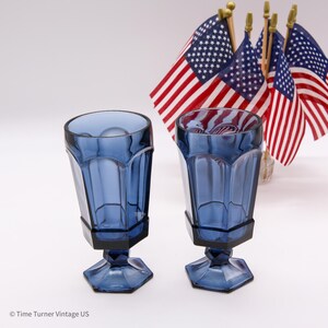 May include: Two matching cobalt blue glass goblets with a faceted, paneled design and a hexagonal base. The glasses feature a thick stem and a wide rim. In the background, several small American flags are arranged on thin wooden poles.