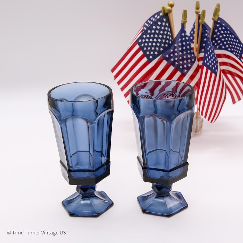 May include: Two matching cobalt blue glass goblets with a faceted, paneled design and a hexagonal base. The glasses feature a thick stem and a wide rim. In the background, several small American flags are arranged on thin wooden poles.