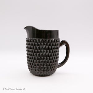 May include: A black glass pitcher with a textured, diamond-like pattern on the lower portion. The pitcher has a smooth, dark rim, a curved handle, and a spout for pouring. The vintage-style pitcher is likely used for serving beverages.