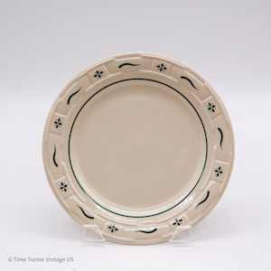 May include: A cream-colored ceramic plate with a green floral and curved line design. The plate has a green border and a decorative rim with small green floral accents. The plate is displayed on a clear stand.