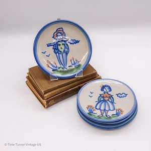 May include: A stack of four small, round ceramic plates with blue rims and hand-painted figures. One plate features a figure with a hat and outstretched arms. The other plates have a figure in a dress. The plates are on a stack of old books.