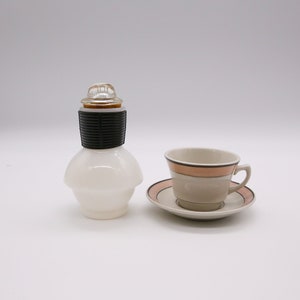 May include: A white glass bottle with a black and silver cap and a white ceramic teacup and saucer with a pink and brown stripe design.