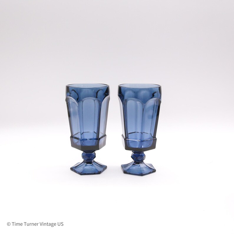May include: A pair of tall, deep blue glass goblets featuring a faceted, hexagonal bowl design and a matching hexagonal base. Each glass has a short, multi-faceted stem connecting the bowl to the base, creating a uniform, structured appearance.
