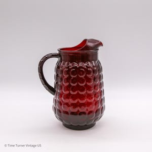 May include: A deep red glass pitcher featuring a prominent hobnail pattern across its rounded body. The vessel includes a curved handle with a textured grip and a wide, molded pouring spout at the top rim, showcasing a translucent finish.