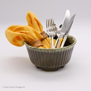 May include: A green ceramic bowl with a yellow napkin and silver cutlery inside. The bowl has a textured, ribbed design.