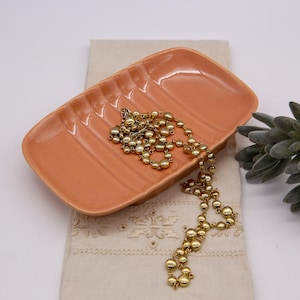 May include: A peach-colored ceramic dish with a ridged design. A gold chain necklace is resting on the dish.
