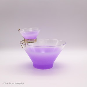 May include: A two-tiered, clear glass serving set with a purple gradient. The smaller bowl is held above the larger one by a gold-colored metal frame. The set is designed for serving appetizers or cocktails.