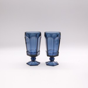 May include: Two blue-tinted drinking glasses. Each glass has a faceted, rectangular body and sits on a hexagonal base. The stem is a short, rounded shape. The glasses are made of a transparent material.