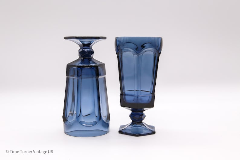 May include: Two deep blue glass goblets with a faceted, paneled design. One goblet stands upright on a hexagonal base with a short, thick stem, while the other is inverted, resting on its flat rim to display the base and stem structure clearly.