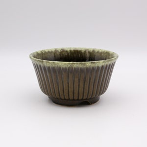 May include: A ceramic bowl featuring a dark brown exterior with vertical ribbed detailing and a contrasting light green glaze around the top rim. The interior of the bowl is dark brown, and the base has a small footed design for stability.