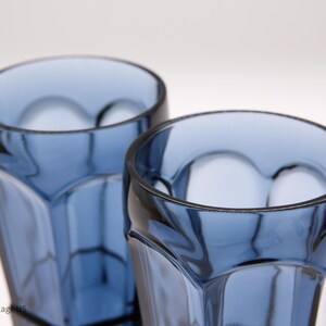 May include: Two blue glass tumblers with a faceted, geometric design featuring vertical panels and rounded arches near the rim. The glasses are transparent with a deep blue hue, showing a clear interior and a smooth, polished top edge.