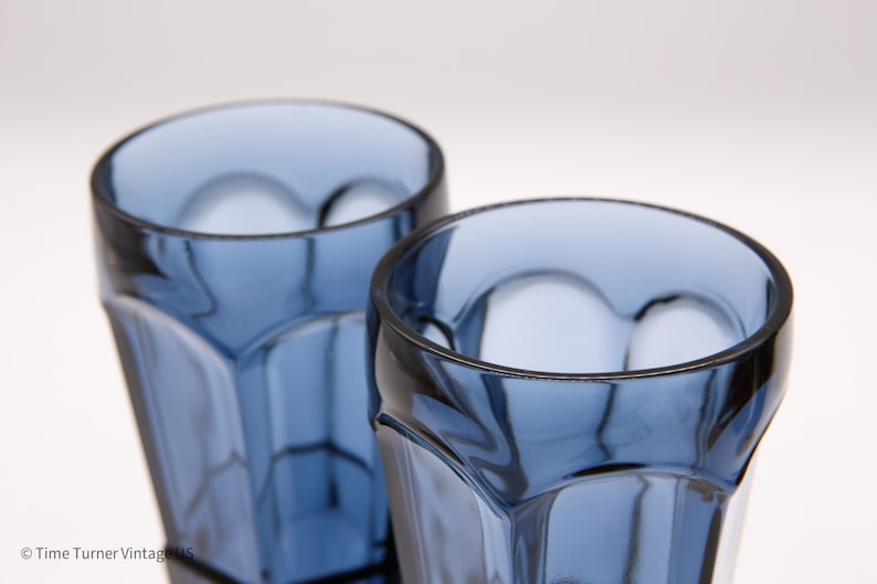 May include: Two blue glass tumblers with a faceted, geometric design featuring vertical panels and rounded arches near the rim. The glasses are transparent with a deep blue hue, showing a clear interior and a smooth, polished top edge.