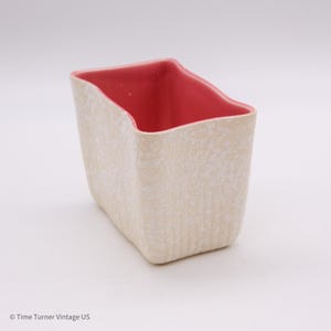 May include: A rectangular, off-white ceramic planter with a textured surface and a red interior. The planter has a unique, asymmetrical shape, with a slightly wavy top edge. The overall design is minimalist and modern.