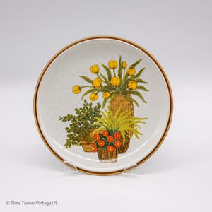 May include: Round ceramic plate with a speckled off-white background and a brown rim. The plate features a floral design with yellow tulips, green foliage, and orange flowers in woven baskets. A vintage decorative piece.