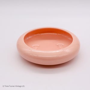 May include: A round, light pink ceramic bowl with a smooth, glossy finish. The bowl has a shallow interior and a wide, rounded rim. The interior of the bowl is a slightly darker shade of pink than the exterior. The bowl is a vintage piece.